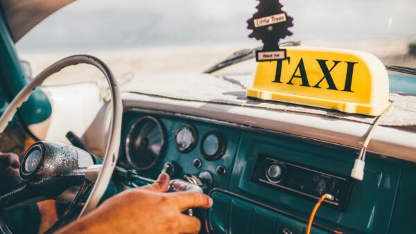 Close-up of a person’s hands on the steering wheel and gear shift in an old car with a yellow TAXI sign on the dashboard and an air freshener shaped like a tree.