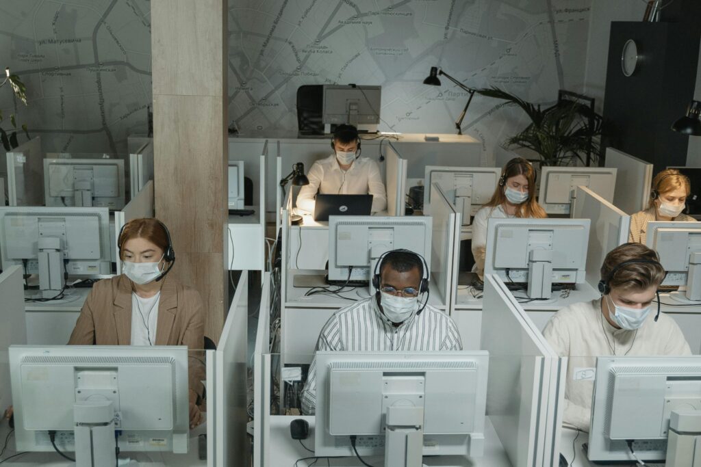 Several people wearing face masks work at computers in separate cubicles in an office with low lighting. A map design is visible on the wall behind them, and they appear focused on their screens.